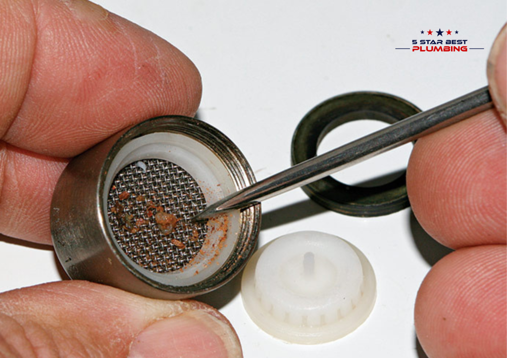 Close-up of a person using a thin metal tool to remove sediment and debris from a stainless steel faucet aerator mesh screen.