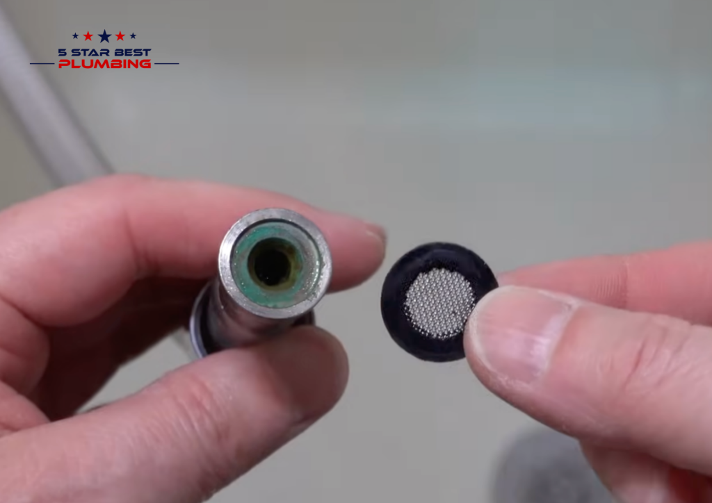 A close-up of a person holding a dirty rubber sealing washer with a metal mesh screen next to an open shower head pipe showing green mineral buildup and oxidation.